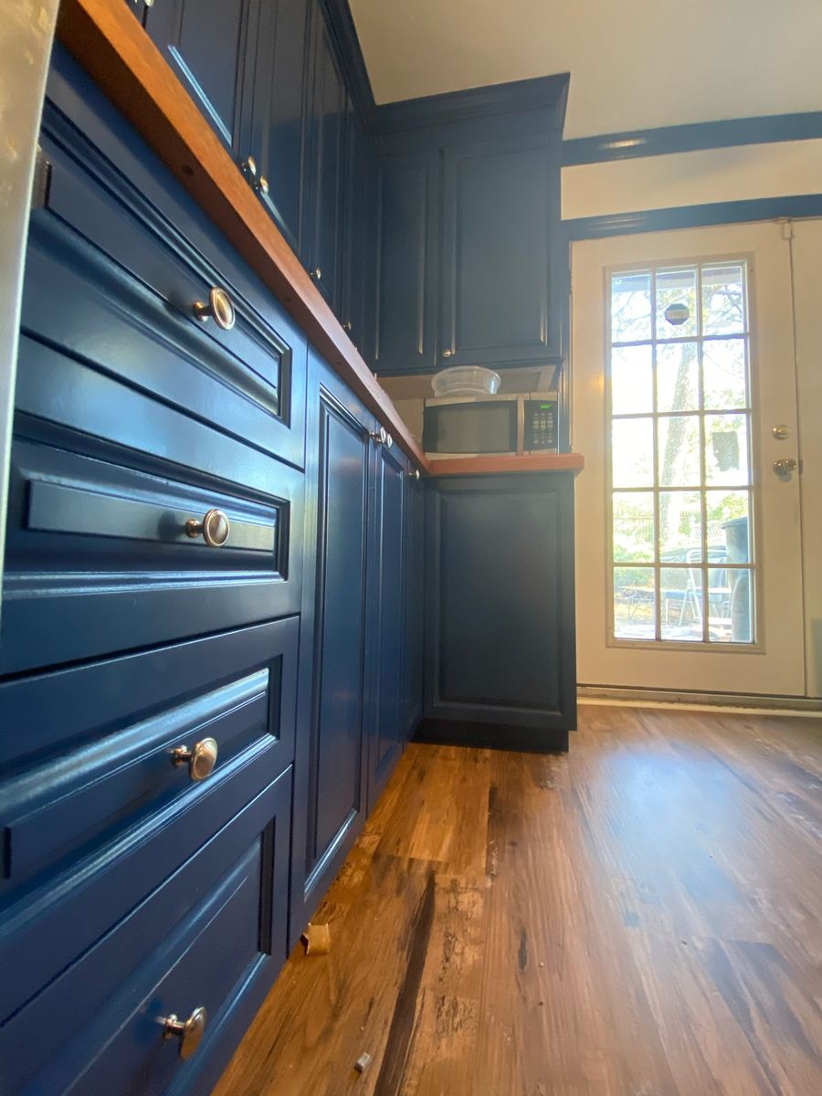 A run of navy drawer bases with brass knobs under a wood counter, navy tall cabinets beyond and a French door at the end