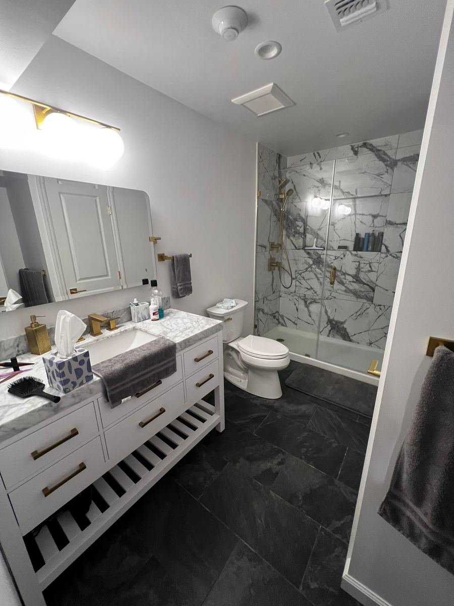 A basement bathroom: a white vanity with gold pulls and an open slatted shelf, and a shower in white veined slab tile behind glass