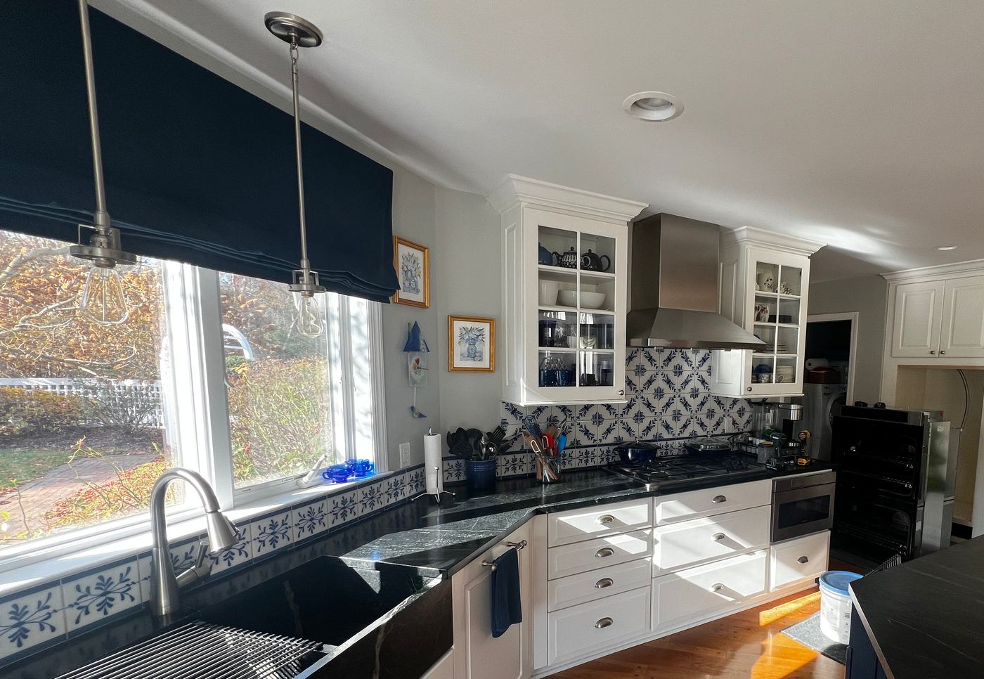 White kitchen with glass-front upper cabinets on both sides of a stainless hood, blue-and-white patterned tile, dark counters and an apron-front sink under a wide window