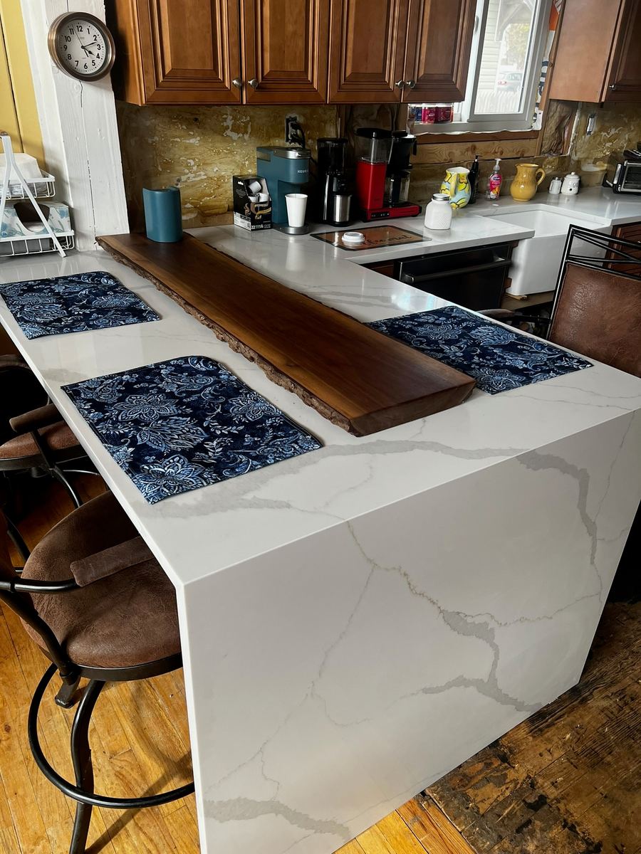 A white waterfall peninsula with gray veining and a dark live-edge wood slab set down its center as a raised bar top, stools beside it