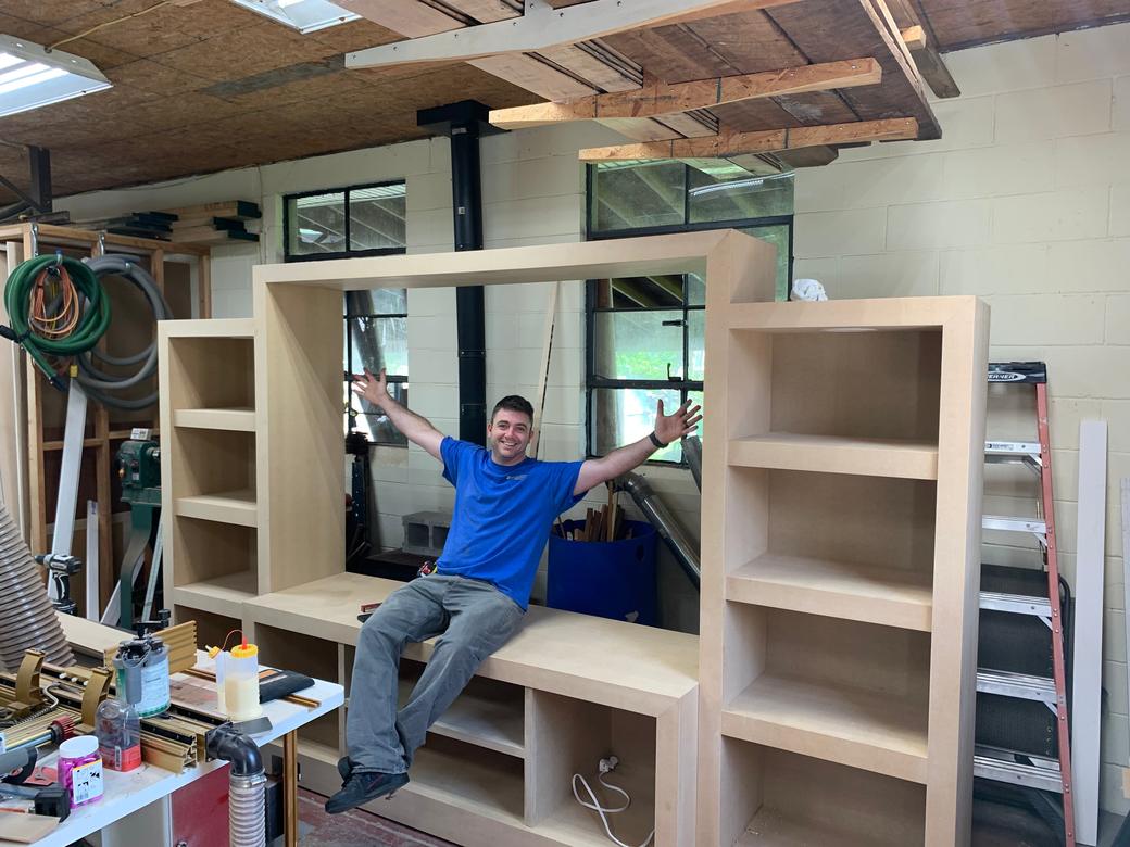 Unfinished entertainment wall standing in a workshop, a man sitting in its open center with his arms out